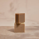 Apeiron Fuß- & Hornhaut Peeling-Seife auf einer Steinfläche mit sanften Pflanzenschatten.