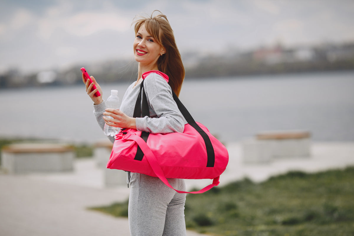Lächelnde Frau in Sportkleidung mit pinker Sporttasche und Wasserflasche im Freien