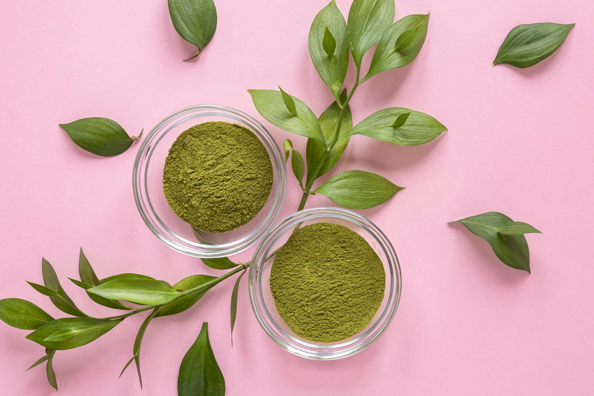 Zwei Glasschalen mit grünem Superfood-Pulver auf rosa Hintergrund mit grünen Blättern.