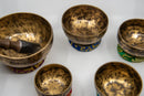 Fünf strukturierte, goldene Klangschalen unterschiedlicher Größe, teils mit tibetischer Schrift und auf farbigen Kissen, angeordnet auf einer weißen Oberfläche, mit einem sichtbaren Holzklöppel.