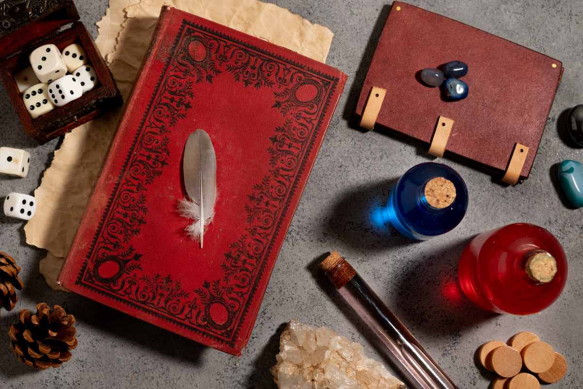 Altes rotes Buch mit Feder, Würfel, Edelsteine und Glasflaschen auf grauem Untergrund.