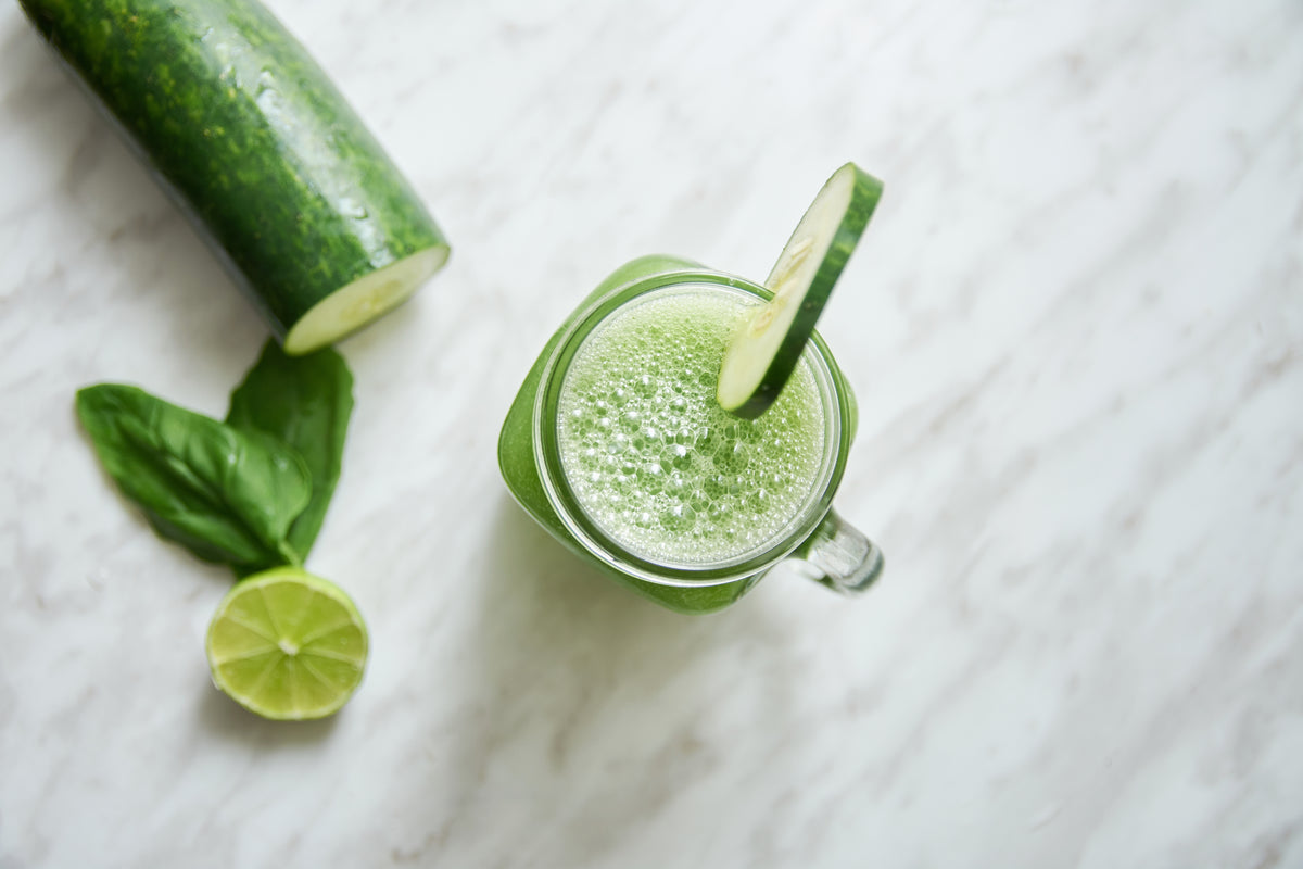 Frischer grüner Smoothie im Glas mit Gurkenscheibe, Limette und Basilikum auf Marmor.