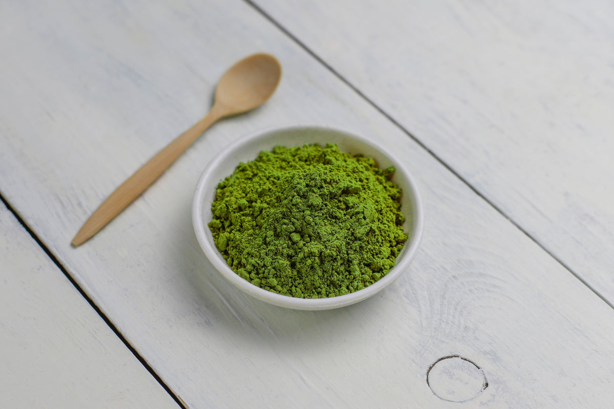 Grünes Matcha-Pulver in einer weißen Schale mit Holzlöffel auf weißem Holztisch.