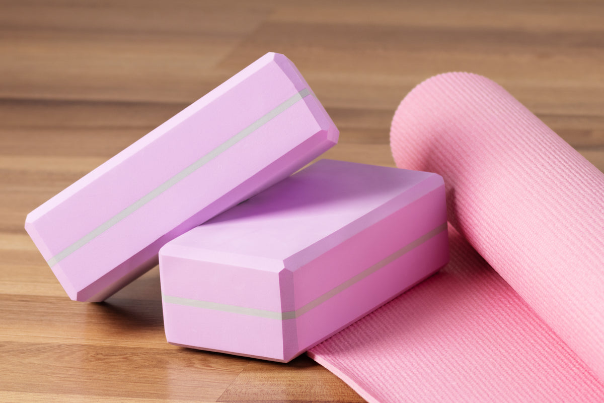 Rosa Yoga-Zubehör: Zwei Yoga-Blöcke und eine gerollte Yogamatte auf Holzboden.