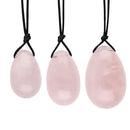 Ein Set aus drei polierten Yoni-Eiern aus Rosenquarz in den Größen klein, mittel und groß. Jedes Ei ist durchbohrt und mit einer schwarzen Rückholkordel versehen, die vertikal aufgehängt gezeigt wird.