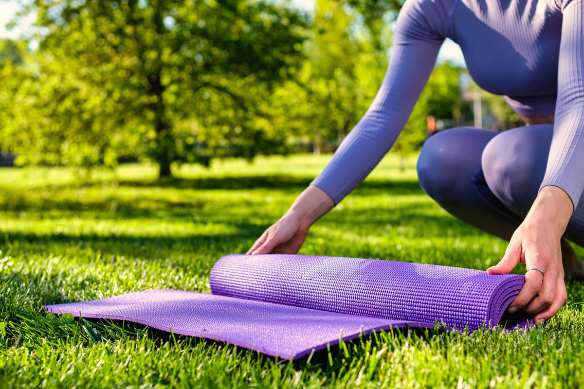 Person rollt eine lila Yogamatte auf einer grünen Wiese im Park aus.